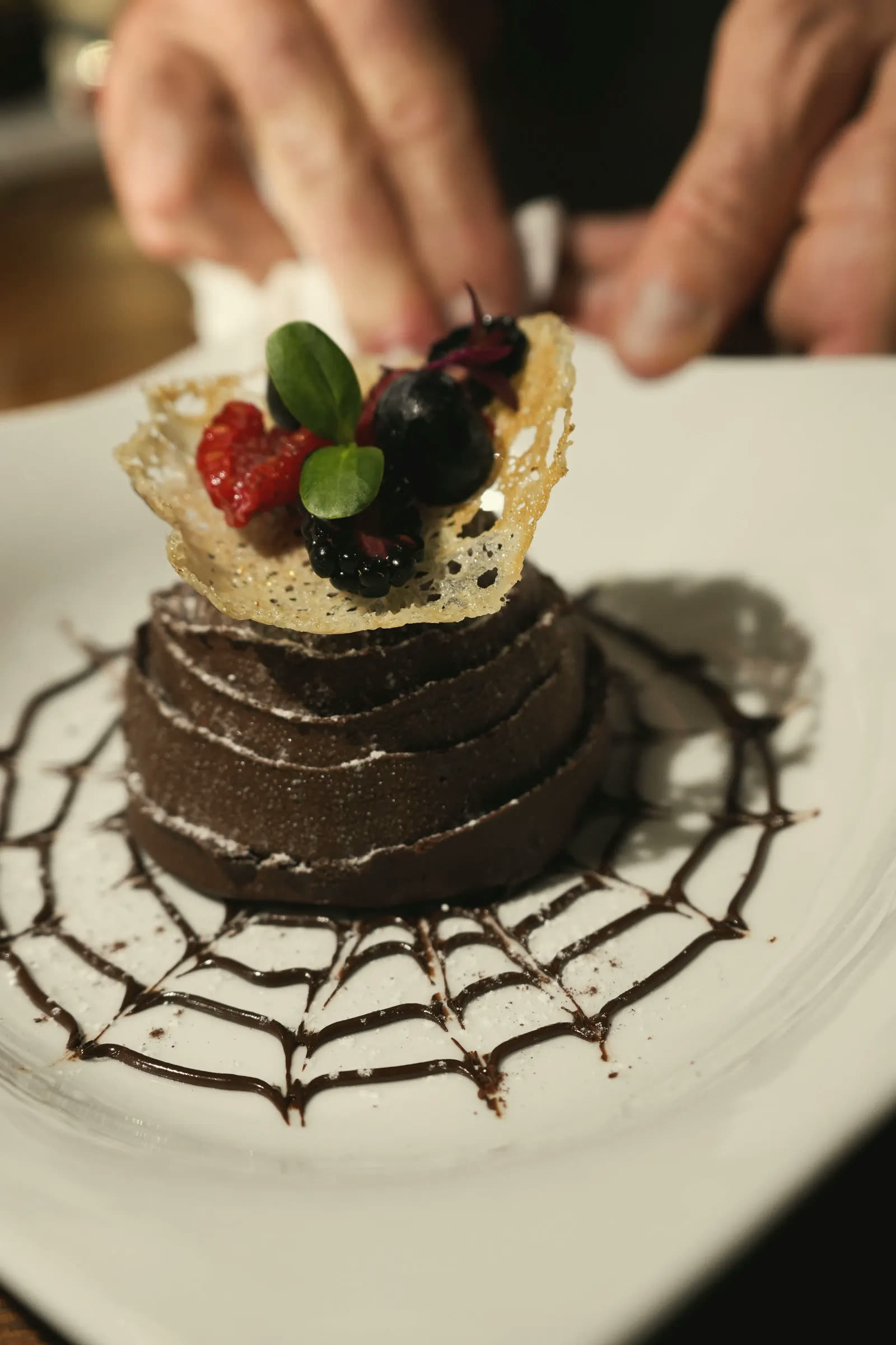 The chef’s hand setting a lace tuile and berries on a chocolate tower