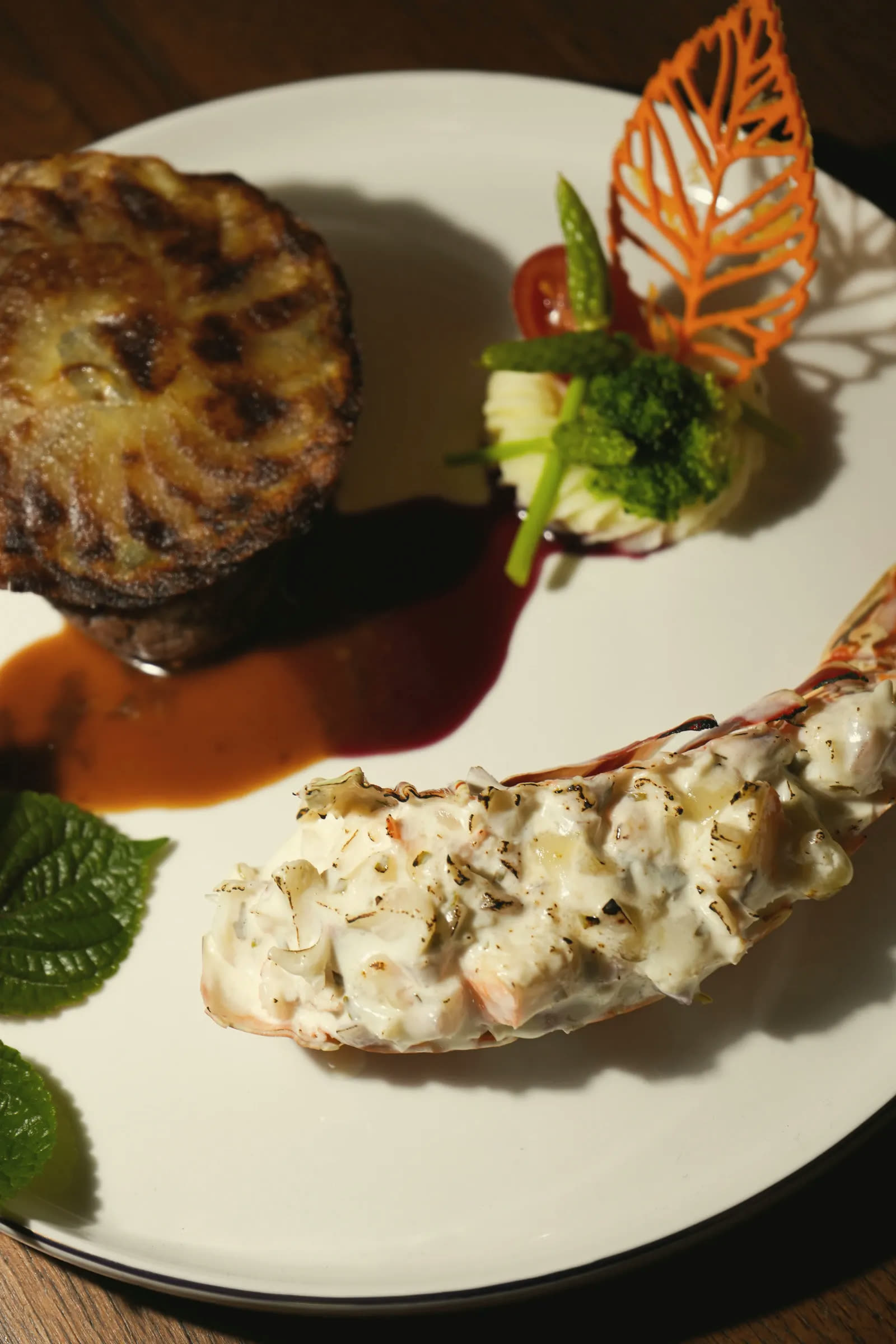 A gratinated lobster tail beside a potato gratin, a carved carrot leaf and two sauces