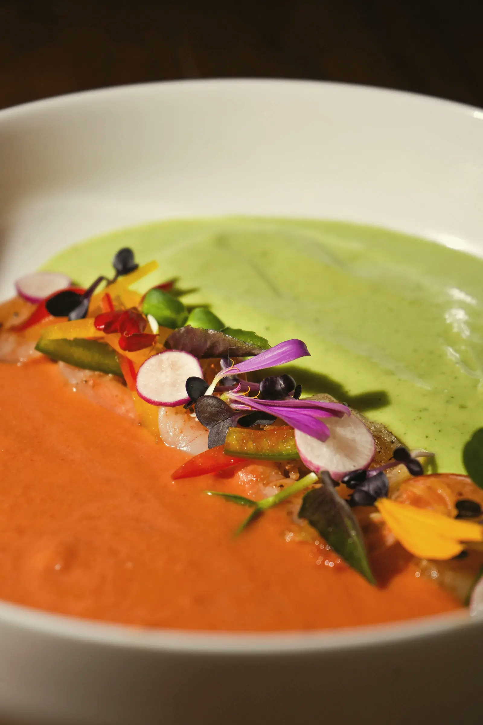 Two soups side by side, green and orange, under crisp vegetables and flowers